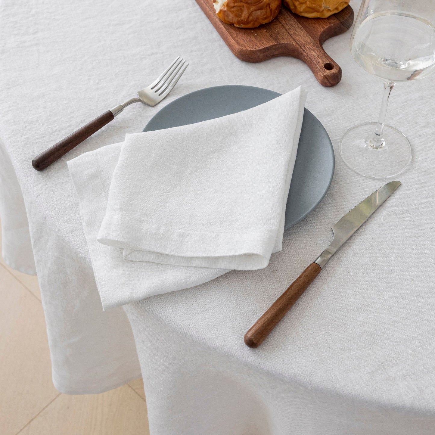 A folded white linen napkin on a gray plate, styled on a white tablecloth with a wooden-handled fork, matching knife, a blue plate, a wine glass, and a wooden cutting board in the background.