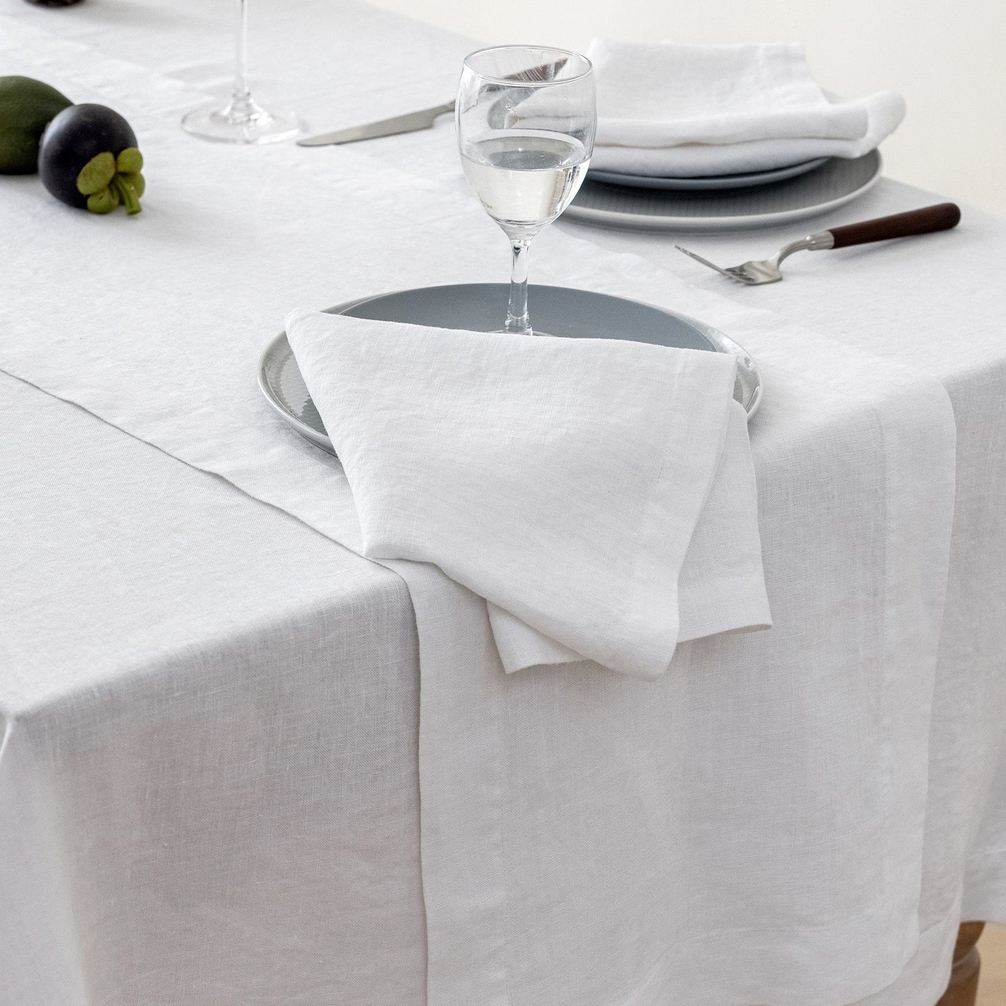 A white linen table runner draped over a white tablecloth, styled with a place setting featuring a white napkin, gray plates, a wine glass, and a fork.