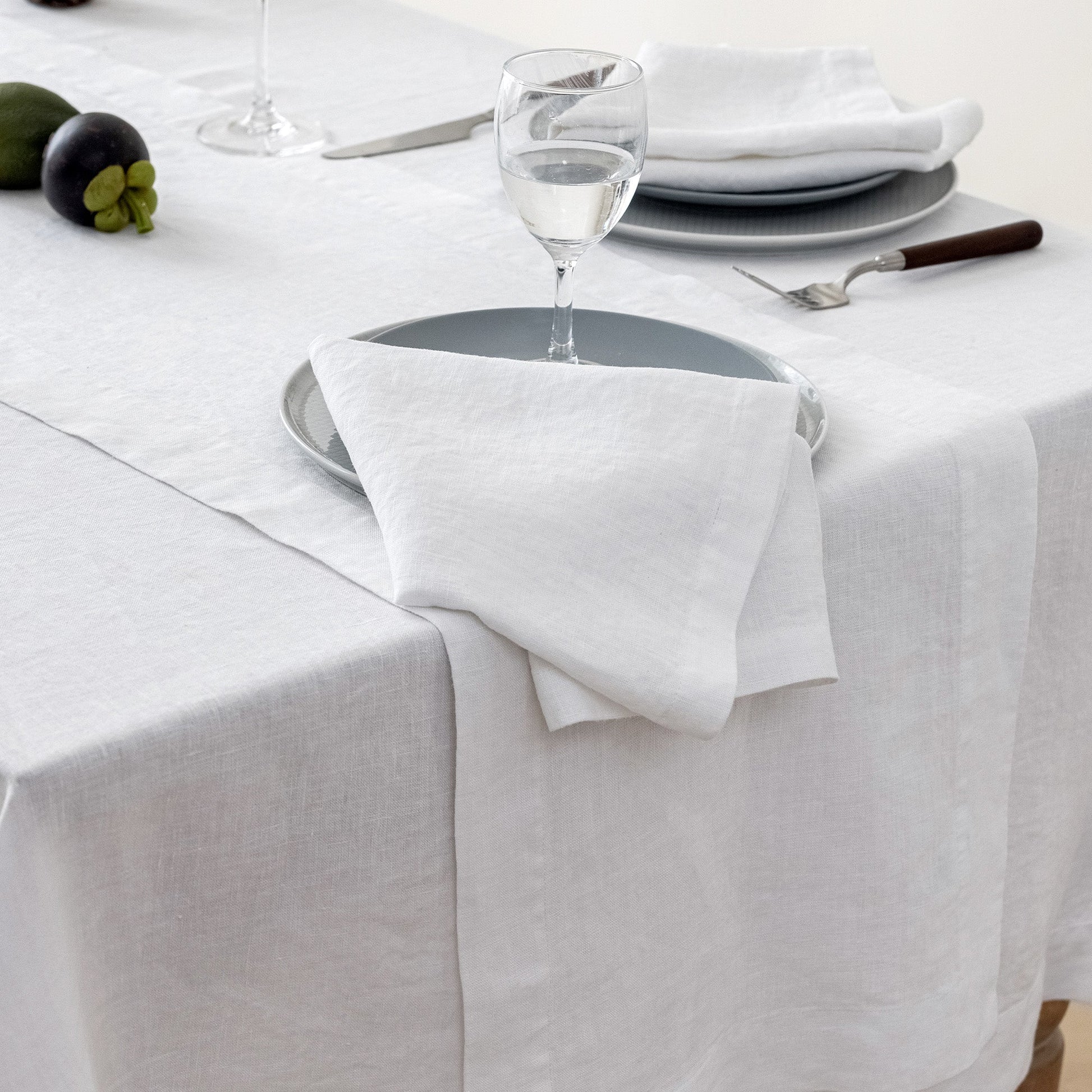 A white linen table runner draped over a white tablecloth, styled with a place setting featuring a white napkin, gray plates, a wine glass, and a fork.