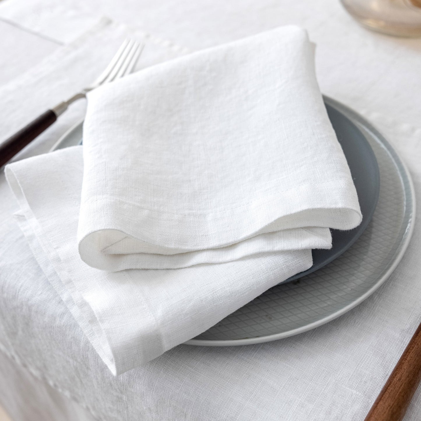 White folded napkin on a stack of gray plates with a fork on a white tablecloth.