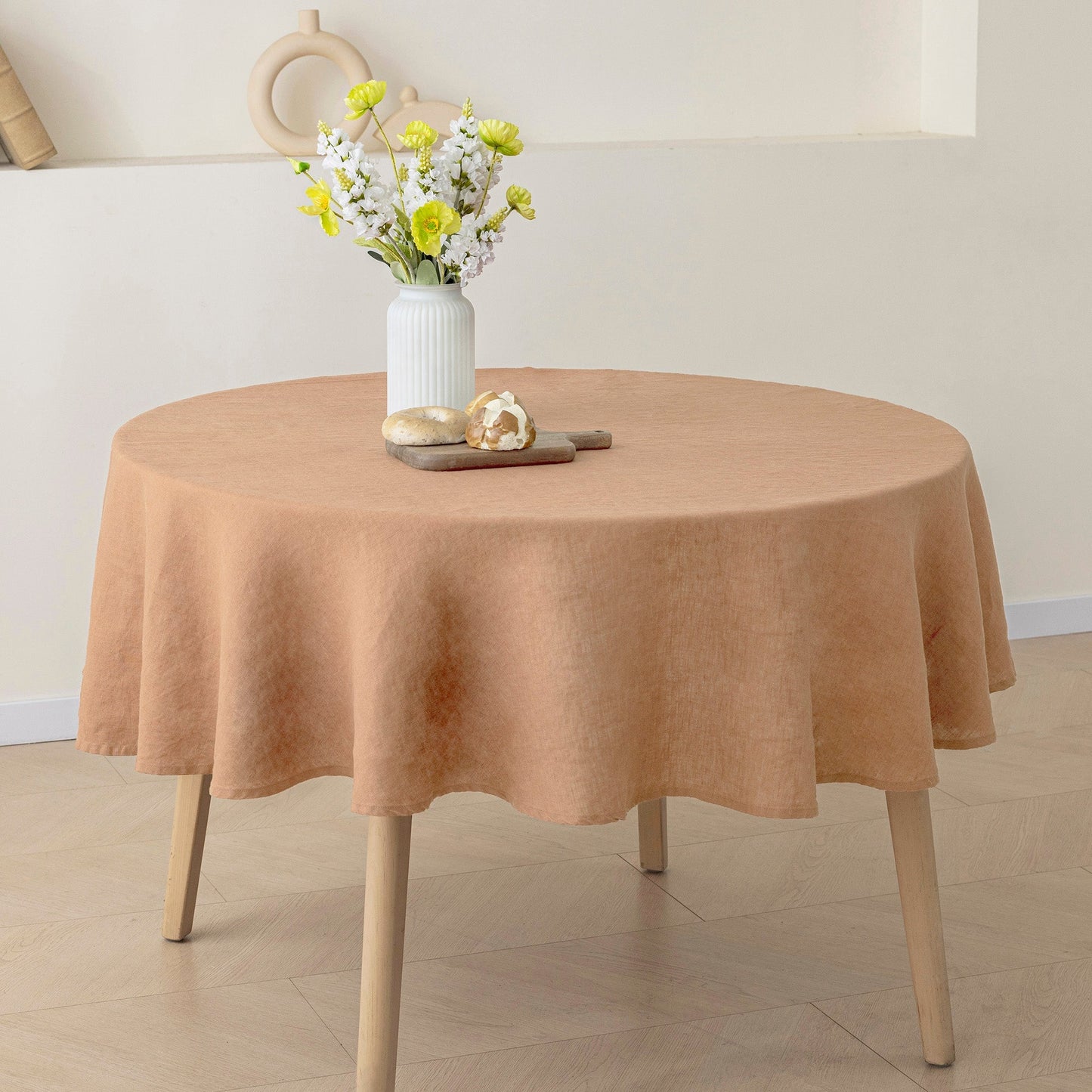 A round table covered with a peach-colored tablecloth, styled with a white vase of yellow and white flowers and a small wooden tray on top. The table has light wood legs and is set against a neutral cream-colored wall and floor.