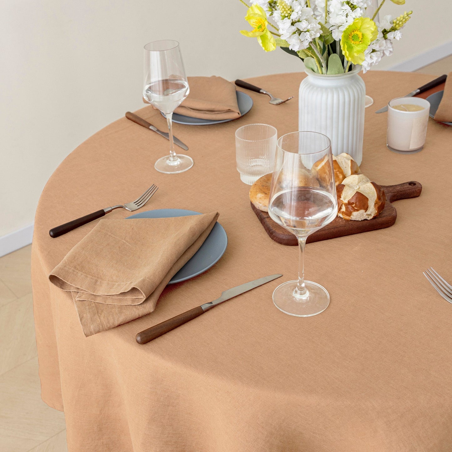 A round table covered with a warm beige tablecloth, styled with blue plates, brown cutlery, wine glasses, a white vase with yellow and white flowers, and bread on a wooden board.