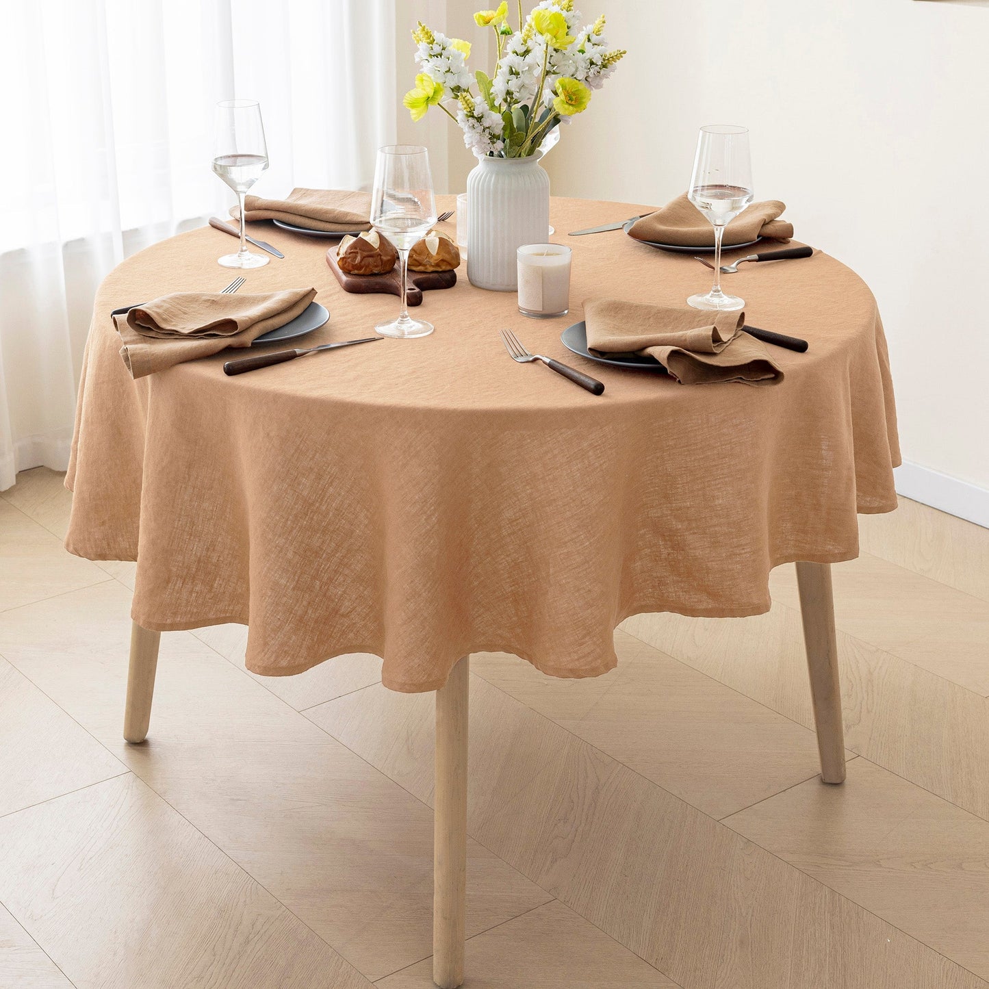 A round dining table covered with a draped peach-beige tablecloth, set with place settings, wine glasses, a vase of flowers, and a candle in a bright room.