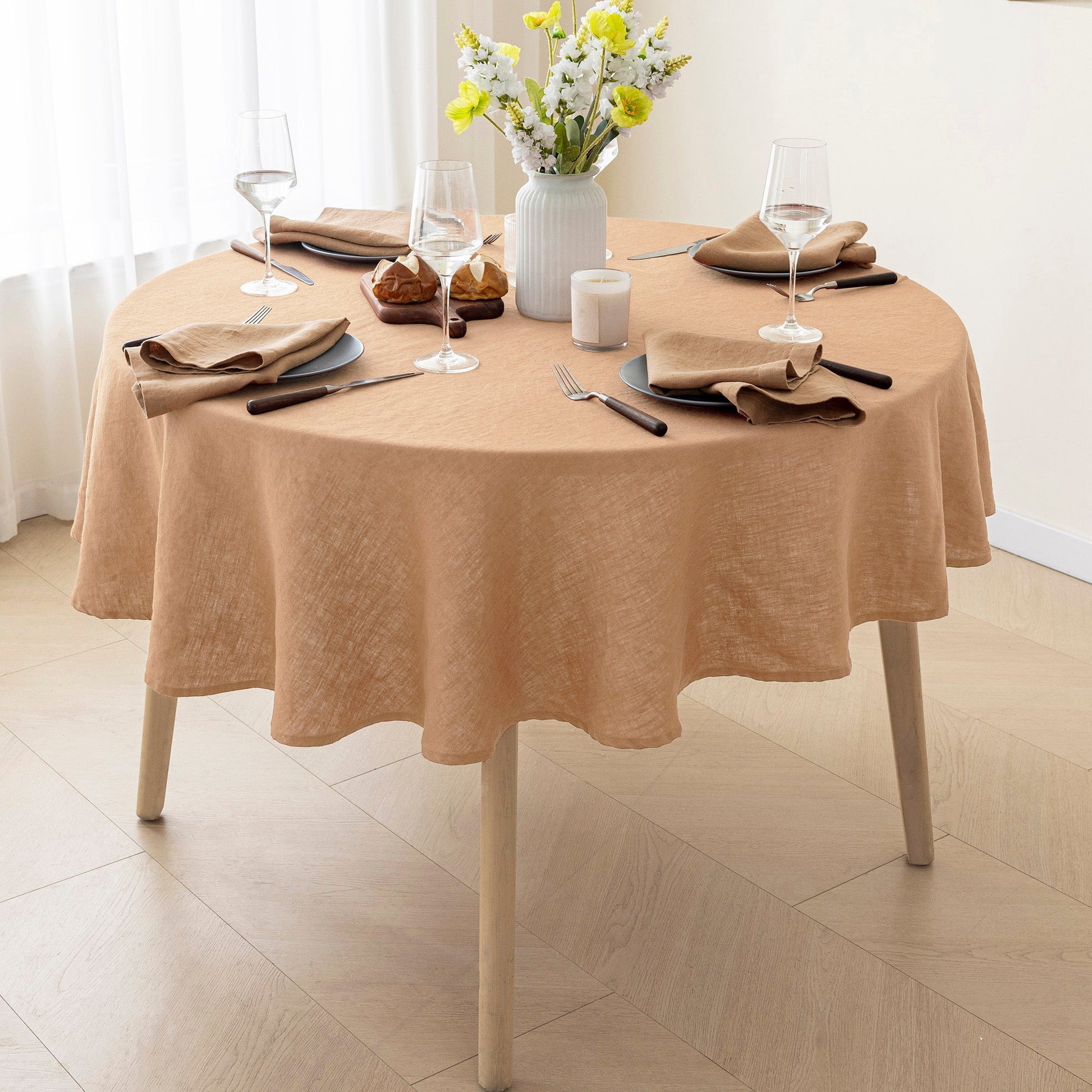 A round dining table covered with a draped peach-beige tablecloth, set with place settings, wine glasses, a vase of flowers, and a candle in a bright room.
