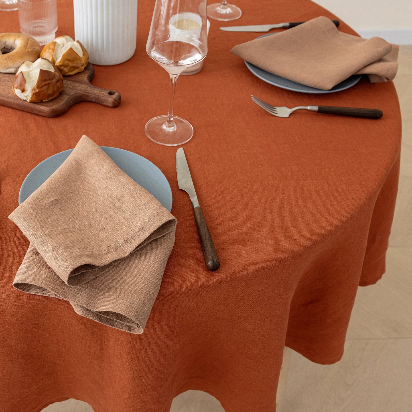 A round table set with a rust-orange tablecloth, light blue plates, beige linen napkins, silver cutlery, and a few pastries on a wooden board.