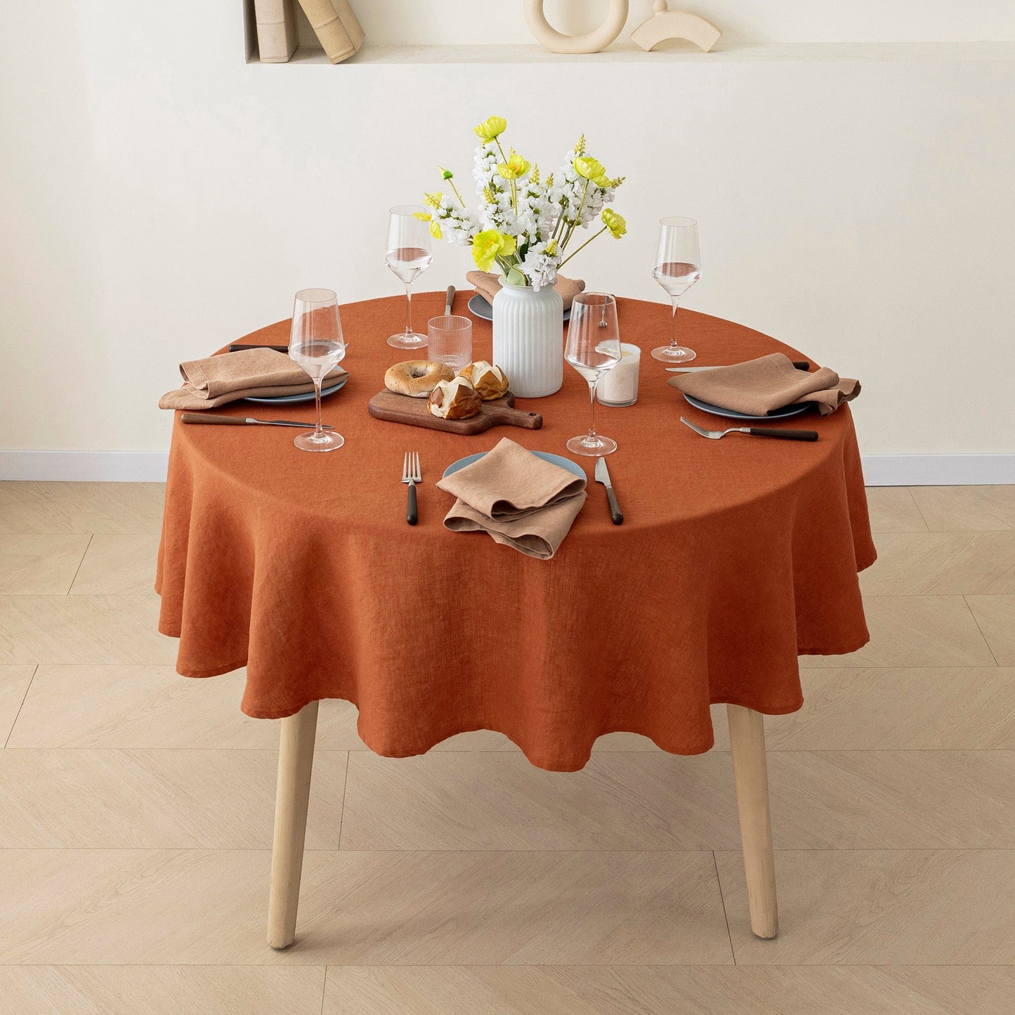 A round dining table covered with a rust-orange tablecloth, styled with beige napkins, clear wine glasses, a white vase with yellow flowers, and a small bread board, set in a bright room with light wood flooring.
