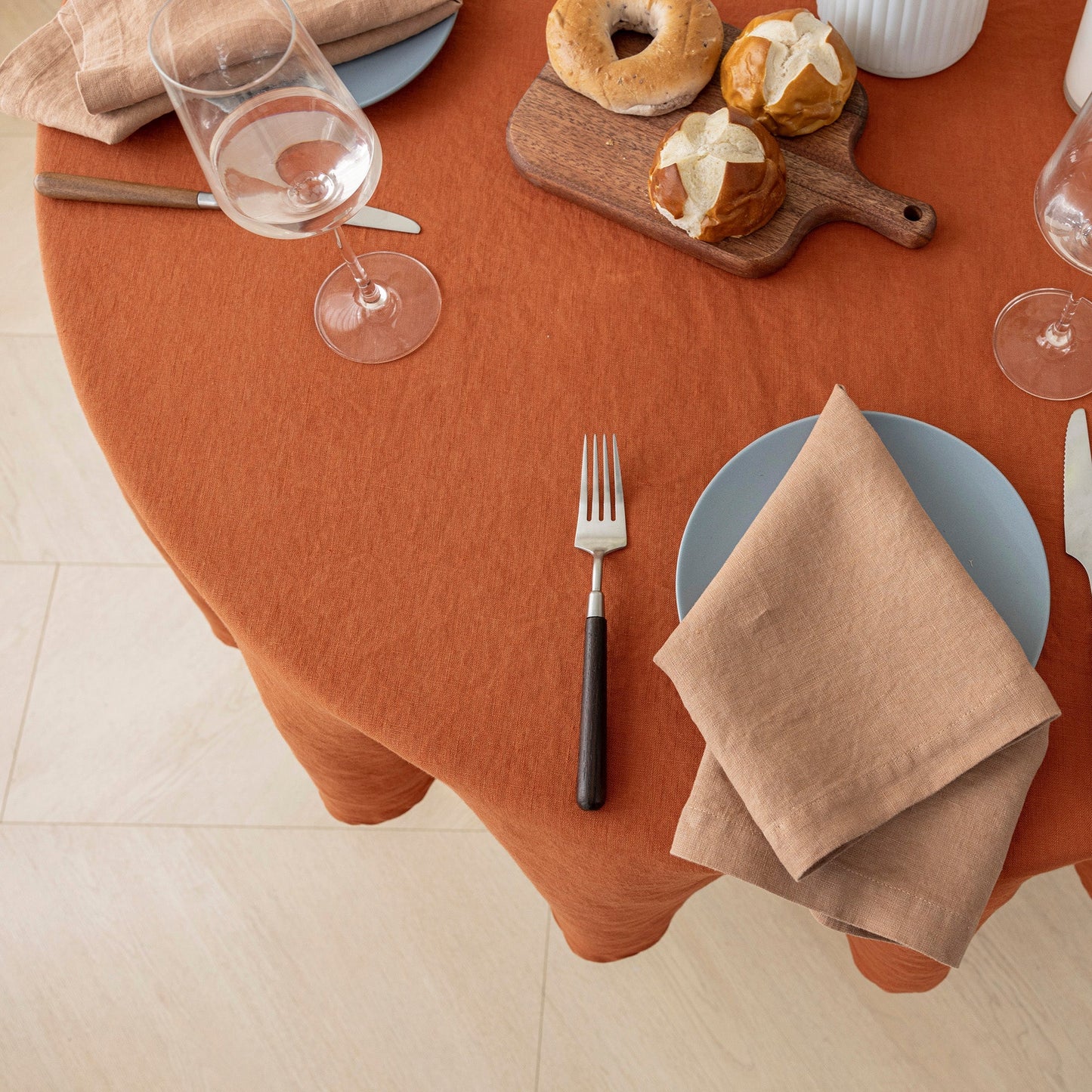 An overhead view of a round dining table covered with a rust-orange tablecloth, styled with a blue plate, beige napkin, fork, wine glasses, and pastries on a wooden board.