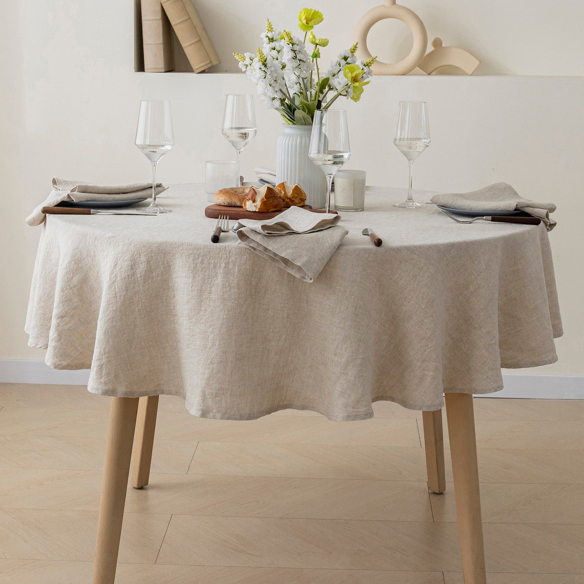 A round dining table covered with a light beige linen tablecloth, set with wine glasses, folded napkins, a small vase of flowers, and bread in a bright, neutral room.