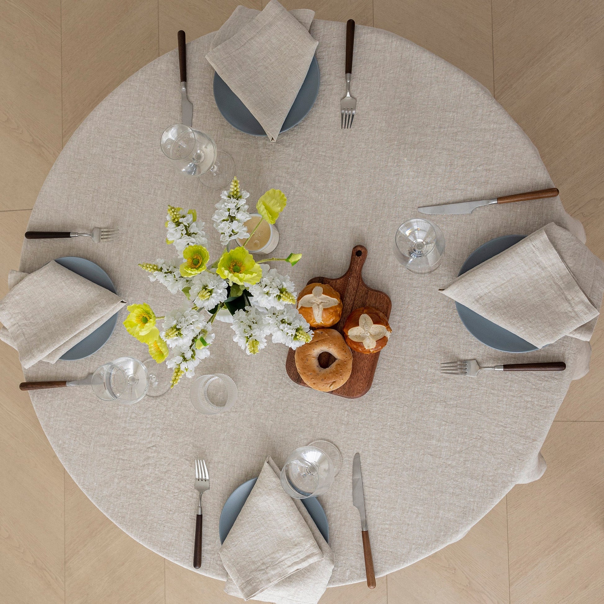 A round dining table set for six with a light beige linen tablecloth, blue plates, folded cream napkins, silverware, a floral centerpiece, and a wooden cutting board with bread on a light wood floor.