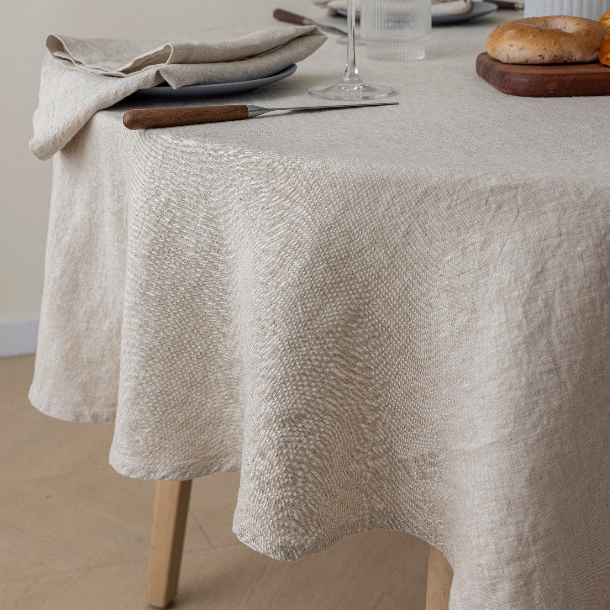 A light beige linen tablecloth draped over a wooden dining table, styled with plates, a folded napkin, a glass, and bread in a softly lit indoor setting.