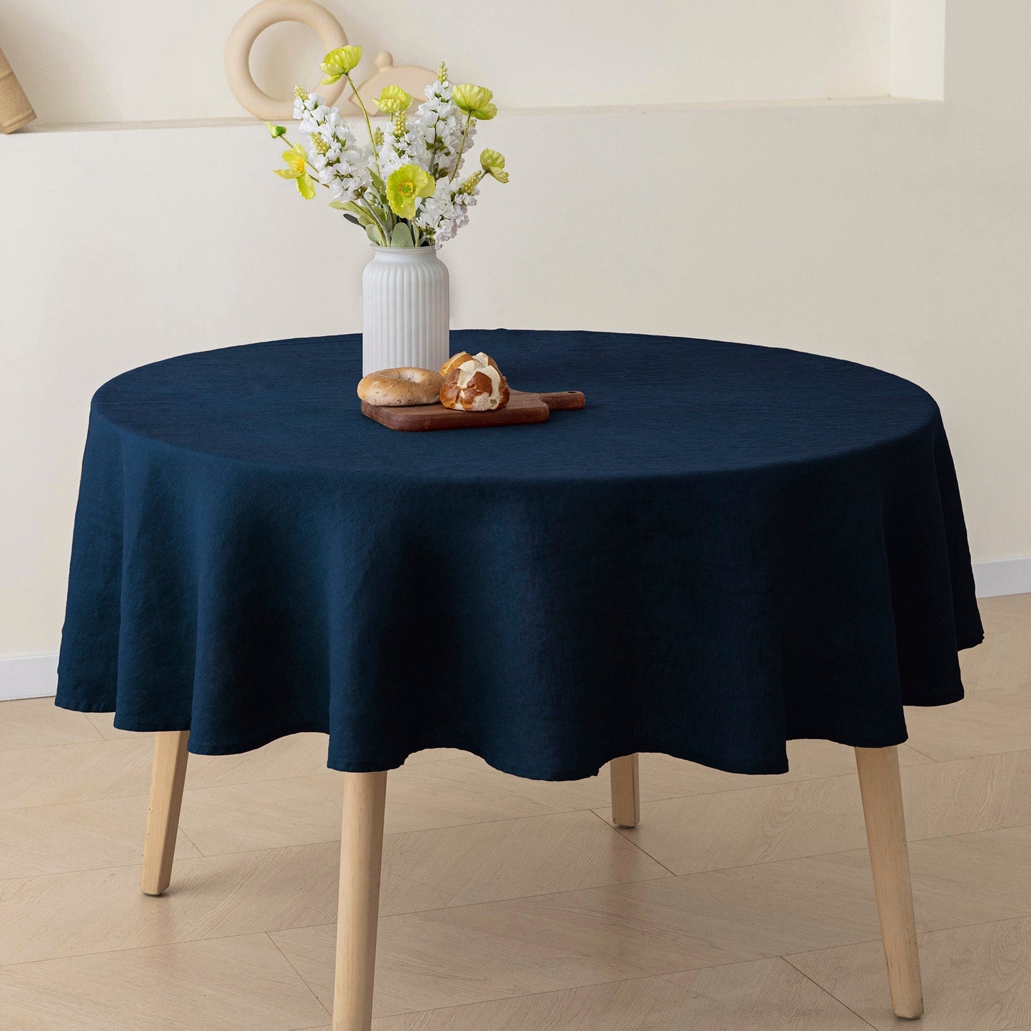 A round table covered with a dark navy blue tablecloth, shown in a styled interior setting with a white vase of flowers and a small pastry on top.
