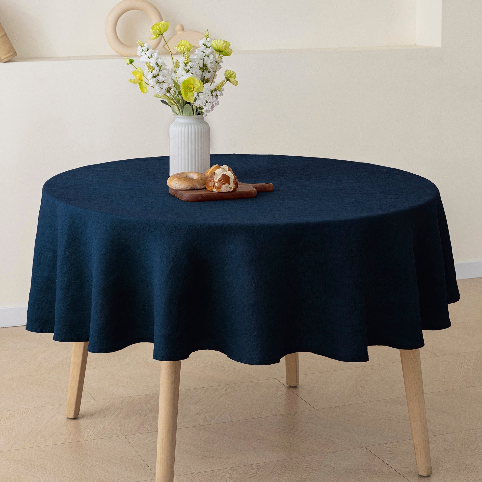 A round table covered with a dark navy blue tablecloth, shown in a styled interior setting with a white vase of flowers and a small pastry on top.