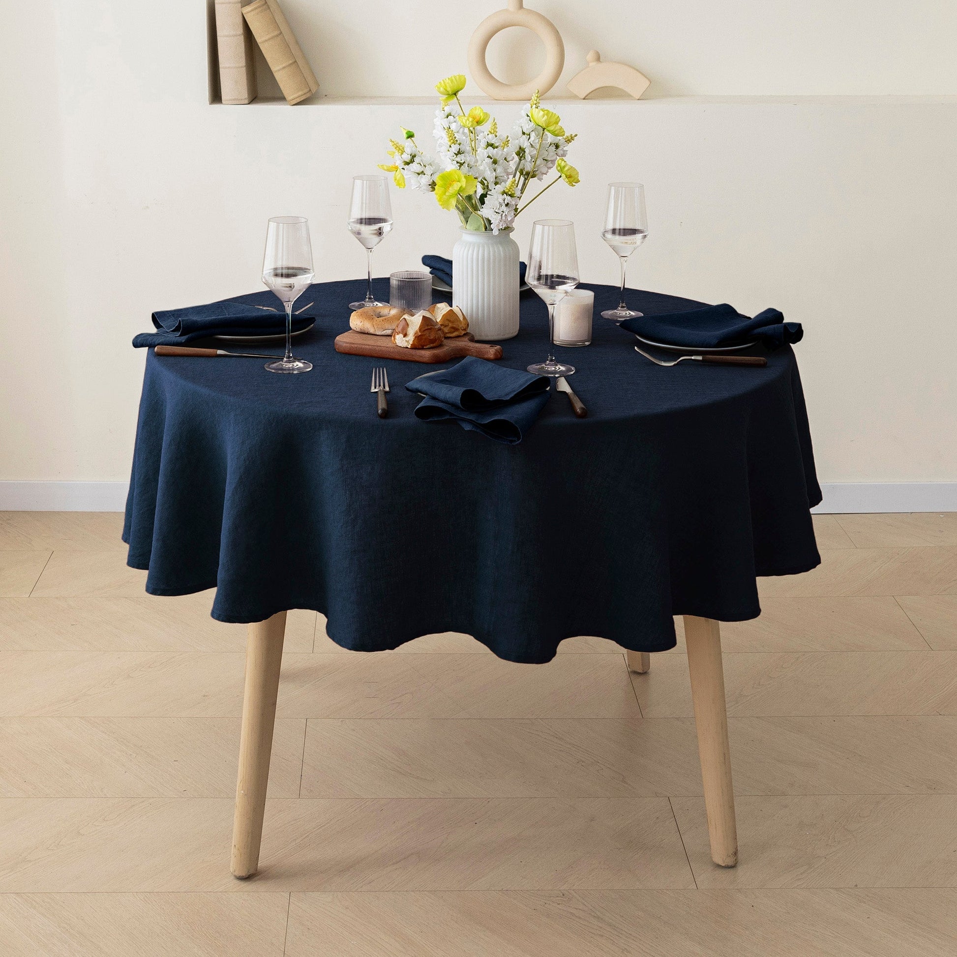 A round table covered with a dark navy blue tablecloth, set with wine glasses, folded navy napkins, bread, and a white vase of yellow flowers in a bright dining room.