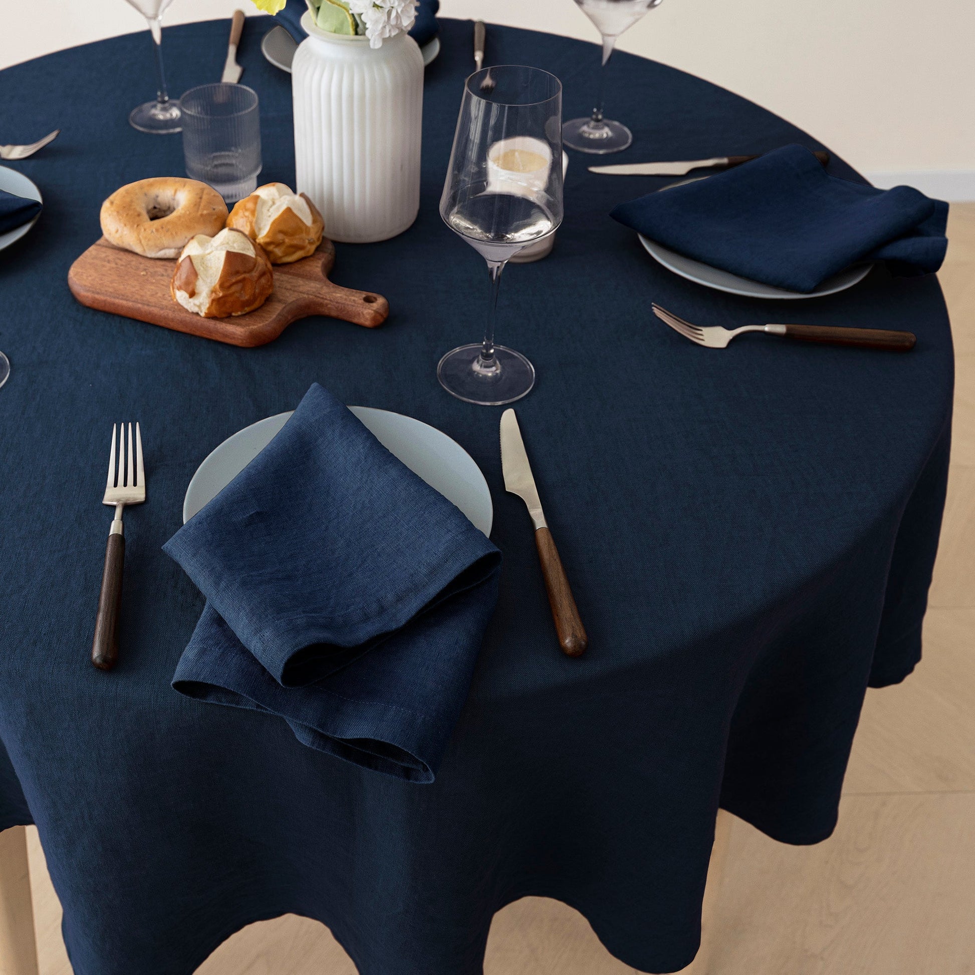 A navy blue tablecloth draped over a round table, styled with navy napkins, white plates, silver and wooden cutlery, wine glasses, a small cutting board with bagels, and a white ribbed vase.