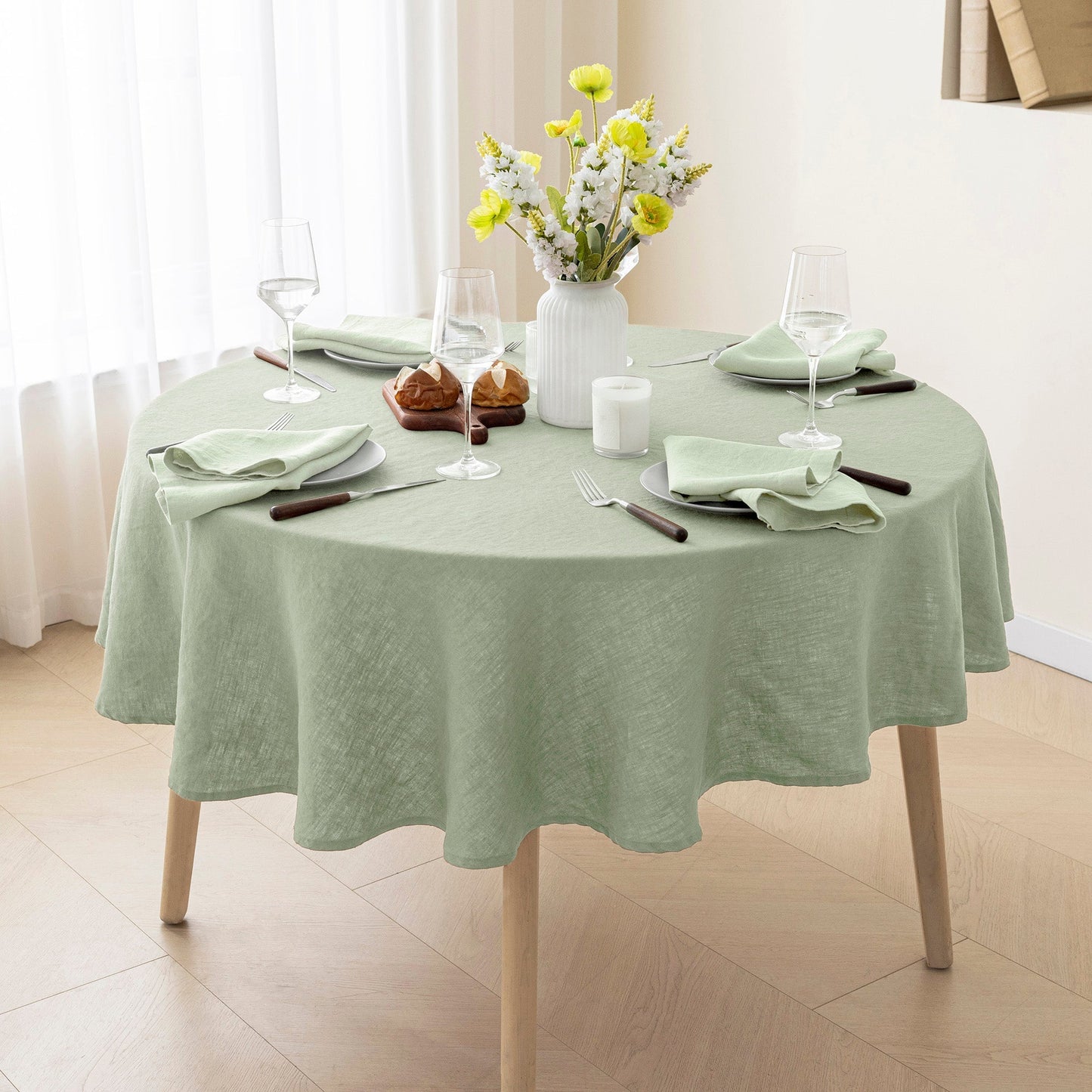 A round dining table covered with a light sage green linen tablecloth, styled with white plates, folded green napkins, wine glasses, a white vase with yellow flowers, and two small brown pastries, set in a bright room with light wood floors and white curtains.