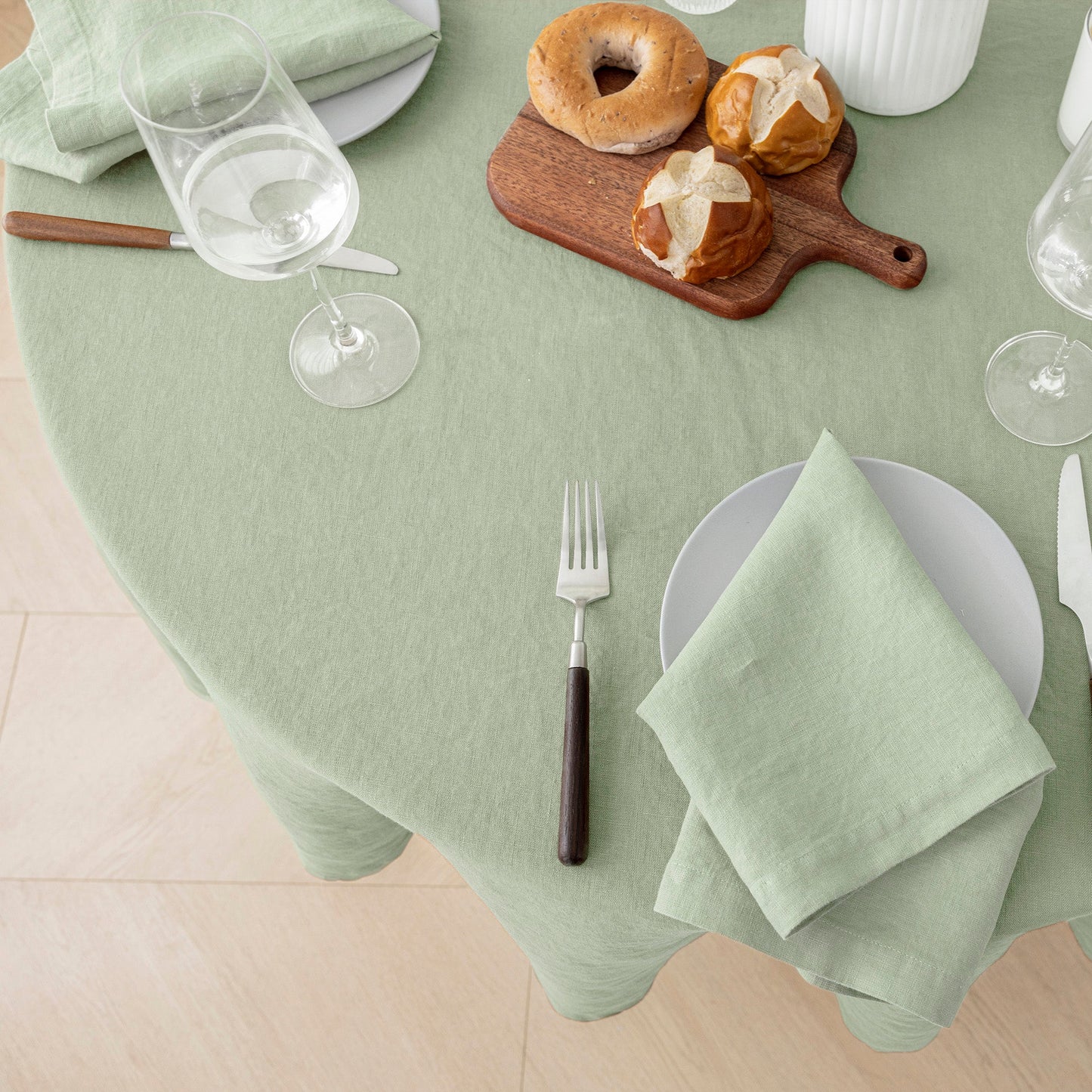 A sage green tablecloth draped over a round dining table, styled with white plates, silver cutlery, wine glasses, a wooden cutting board with bagels, and a folded napkin.