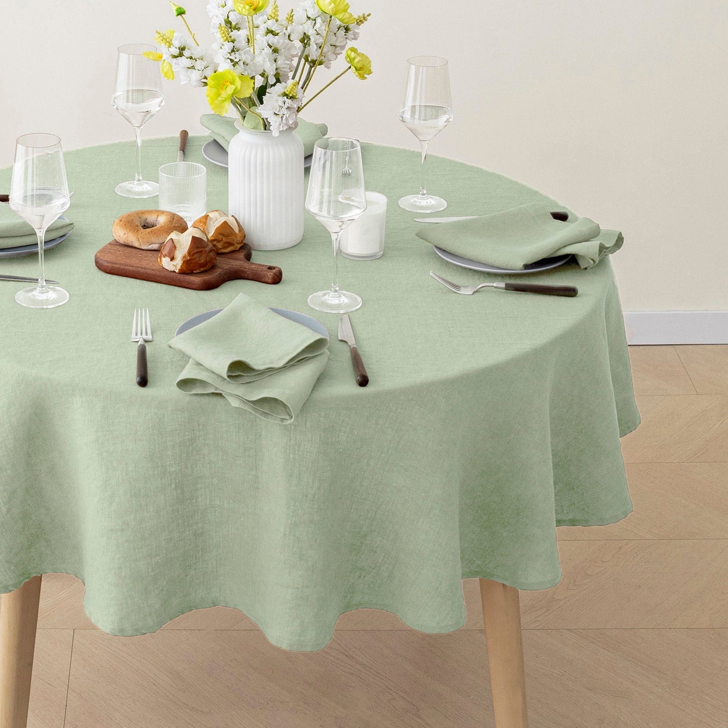 A sage green round tablecloth draped over a dining table, styled with wine glasses, folded napkins, a cutting board with bread, and a white vase of flowers.