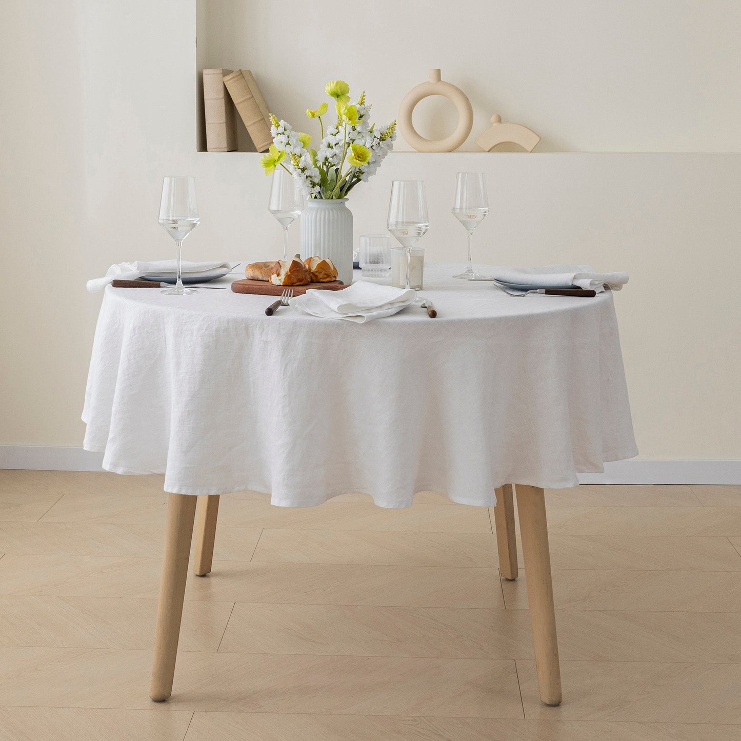 A round dining table covered with a white linen tablecloth, set with wine glasses, plates, napkins, a vase of yellow flowers, and small pastries. The table has light wood legs and is placed in a minimalist room with beige walls and floor.