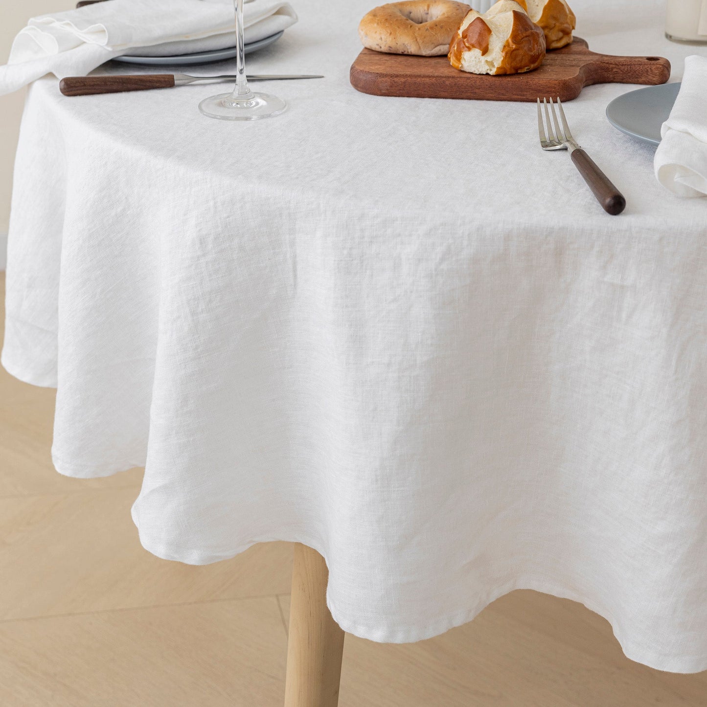 A white round tablecloth draped over a dining table, styled with plates, a wine glass, a wooden cutting board with bagels, and a fork on a light wood floor.
