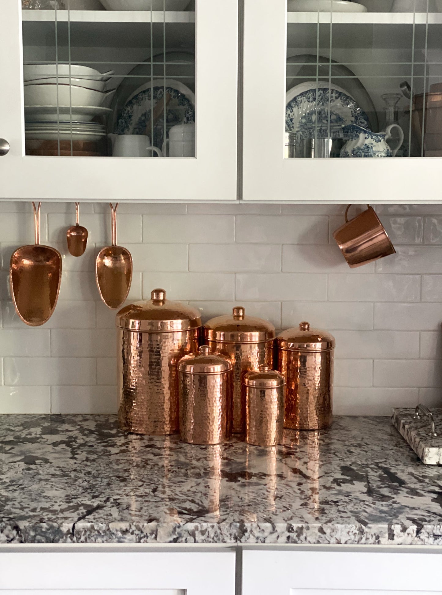 Copper kitchen canisters and utensils on a marble countertop with white cabinets in the background.