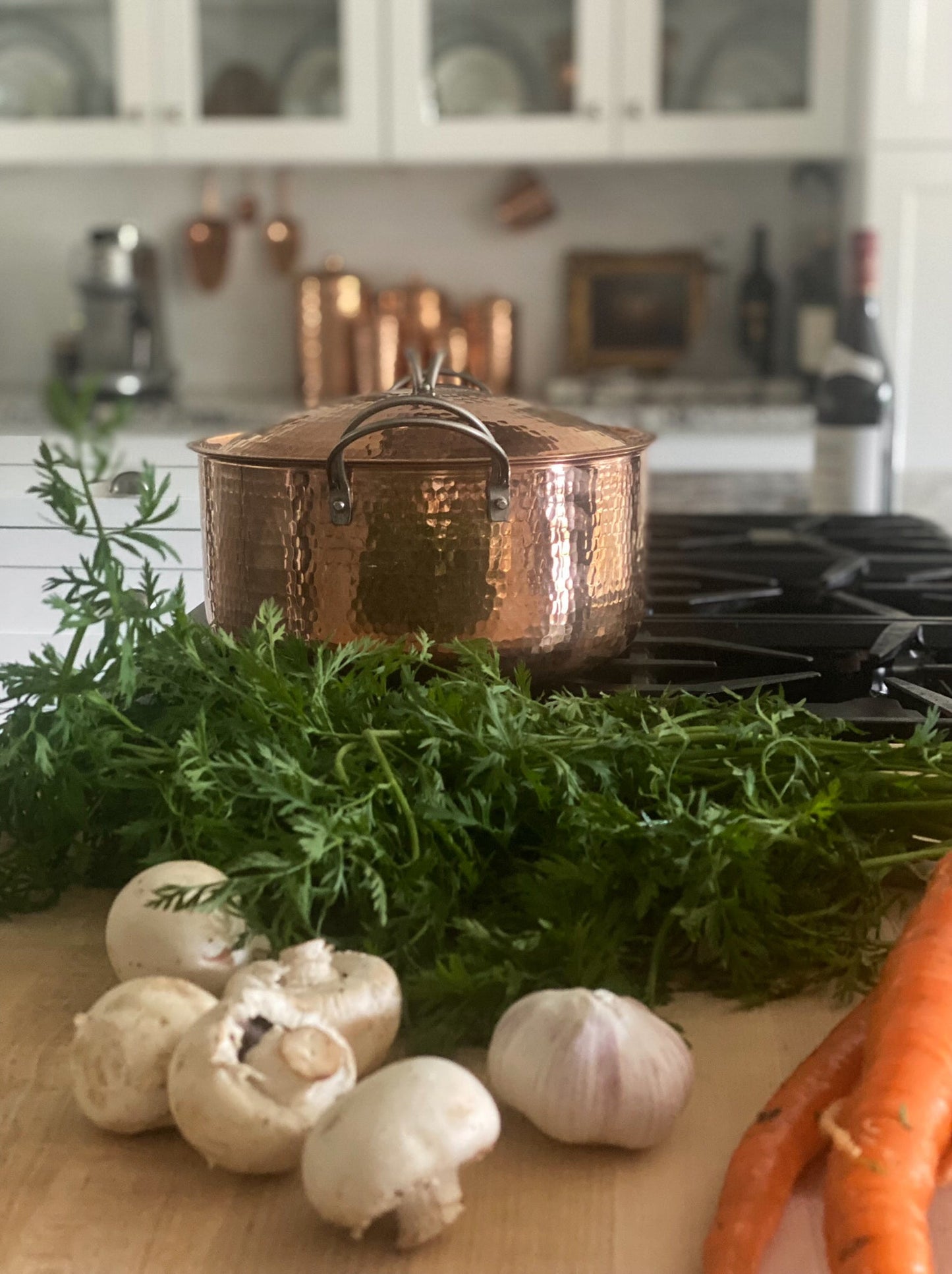 Copper pot on a stove with fresh vegetables and herbs on a wooden surface in a kitchen.