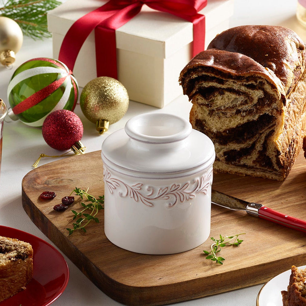 Decorative white ceramic jar on a wooden cutting board with Christmas ornaments and bread in the background.